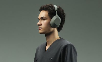Un retrato de perfil de una persona en estudio con el pelo rizado oscuro mirando hacia la izquierda sobre un fondo verde sólido y apagado. Está usando unos audífonos tipo diadema Sony WH-1000XM6 Olive Gray y una chaqueta gris de doble botonadura con botones oscuros.