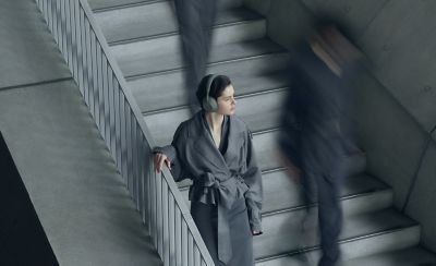Una imagen desde arriba de una persona de pie en una escalera de hormigón mirando hacia la derecha con los audífonos tipo diadema Sony WH-1000XM6 Olive Gray. Está usando una chaqueta gris estilo gabardina. La escena transmite movimiento, con las siluetas desenfocadas de otras personas que suben y bajan las escaleras a su alrededor.