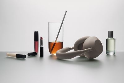 Una escena de naturaleza muerta con un par de audífonos tipo diadema Sony WH-1000XM6 en color Sandstone sobre una superficie gris apagada. A la izquierda de los audífonos, hay un vaso alto y transparente lleno de un líquido color ámbar y una pajita de metal, con junto con un tubo abierto de lápiz labial de color rojo rosáceo, un brillo labial líquido y un tubo de corrector. A la derecha, hay un frasco de perfume de cristal transparente lleno de un líquido verde claro y cubierto con una tapa gris.