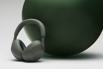 Una toma de primer plano de un par de audífonos tipo diadema Sony WH-1000XM6 en color Sandstone sobre una superficie blanca. Los audífonos están apoyados en una gran esfera mate verde oscuro que domina el lado derecho del cuadro bajo una luz suave.