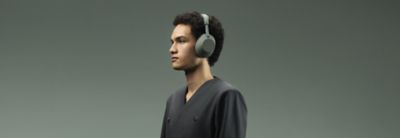Um retrato de perfil de estúdio de uma pessoa com cabelo encaracolado escuro voltado para a esquerda contra um fundo verde sólido suave. Está a usar auscultadores over-ear Sony WH-1000XM6 na cor Olive Gray e um casaco cinzento com dupla fileira de botões escuros.