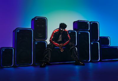 A dynamic wall of the ULT POWER SOUND speakers stacked in varying vertical and horizontal orientations, with each unit outlined by glowing, neon blue LED light rings against a blue and teal gradient background. A person wearing a dark jacket with orange stripes and sunglasses sits on the ULT TOWER MAX speaker in the middle.