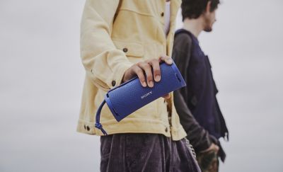 Photograph of a person wearing a pale yellow jacket and holding a blue Sony ULT FIELD 1 portable wireless speaker. Another person stands slightly behind them.