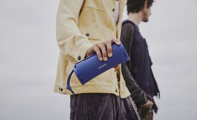 Fotografía de una persona con una chaqueta amarilla claro sosteniendo un parlante inalámbrico portátil ULT FIELD 1 de Sony azul. Hay otra persona parada ligeramente detrás de la primera.