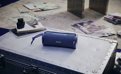 Fotografía de estilo de vida de un parlante inalámbrico portátil ULT FIELD 1 de Sony azul sobre un baúl de avión metálico plateado. En el fondo, hay libros, revistas y artículos de papelería desperdigados por el suelo.