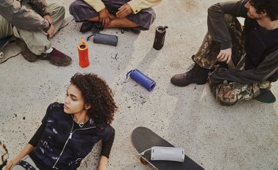 Una fotografía desde un ángulo elevado en la que se muestra a cuatro personas con patinetas sobre el concreto. Hay una persona con cabello rizado oscuro recostada, mientras que otras tres están sentadas en círculo. Hay varios parlantes inalámbricos portátiles ULT FIELD 1 de Sony naranjas, azules, grises y blancos desperdigados por el suelo y sobre una patineta.