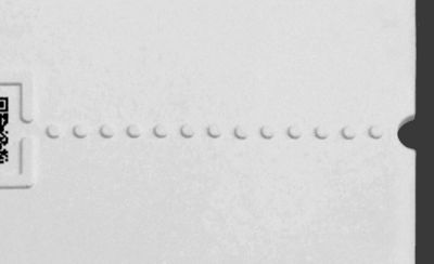 A close-up of a white textured paper surface with a horizontal row of raised tactile dots across the centre. Part of a QR code is visible on the left edge with a raised tactile border around it. A small notch appears on the right edge.