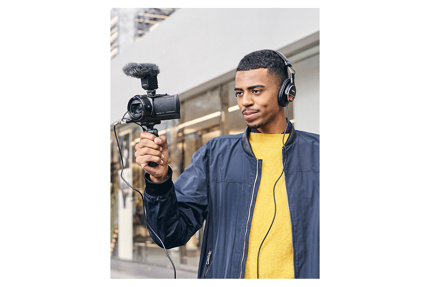 Ein Mann mit Kopfhörern hält eine Kamera von Sony, an der ein Aufnahmegriff und ein Mikrofon befestigt sind.