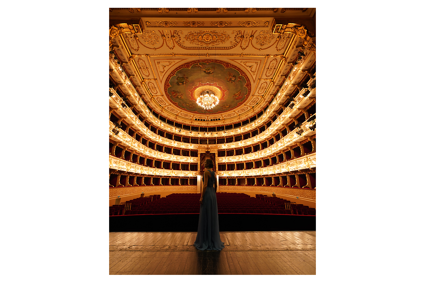 A woman wearing a long blue dress stands on a stage in front of an empty theatre.