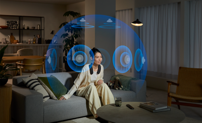 Person sitting on a grey sofa in a living room wearing BRAVIA Theatre U while watching a movie with a blue sphere of phantom speakers all around