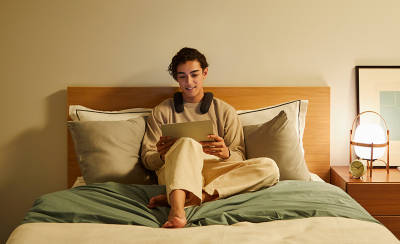 Person sitting on a bed wearing BRAVIA Theatre U while watching content on a tablet with a bedside table to right with lamp and clock