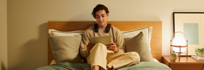 Person sitting on a bed wearing BRAVIA Theatre U while watching a movie on a tablet with wall behind 