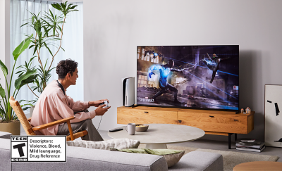 Person sitting on a chair in a living room wearing BRAVIA Theatre U while playing a game on a big screen TV with screenshot of a Spider-Man 2 street scene and Teen ESRB logo bottom left