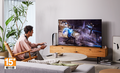 Person sitting on a chair in a living room wearing BRAVIA Theatre U while playing a game on a big screen TV with screenshot of a Spider-Man 2 street scene and 15+ logo bottom left