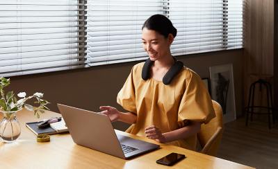 Person sitting at a desk wearing BRAVIA Theatre U on a video call using laptop with notebook and pen on desk to left and smartphone on desk to right 