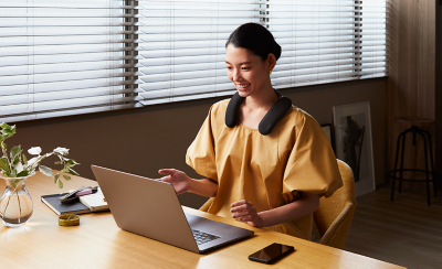 Person sitting at a desk wearing BRAVIA Theatre U on a video call using laptop with notebook and pen on desk to left and smartphone on desk to right