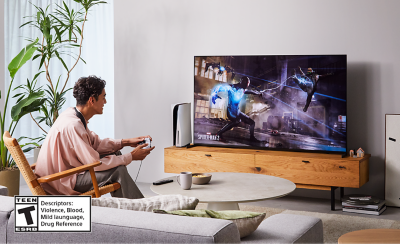 Person sitting on a chair in a living room wearing BRAVIA Theatre U while playing a game on a big screen TV with screenshot of a Spider-Man 2 street scene and Teen ESRB logo bottom left