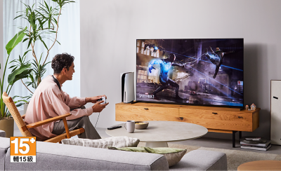 Person sitting on a chair in a living room wearing BRAVIA Theatre U while playing a game on a big screen TV with screenshot of a Spider-Man 2 street scene and 15+ logo bottom left