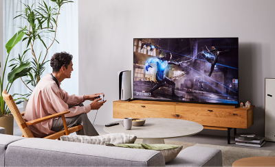 Person sitting on a chair in a living room wearing BRAVIA Theatre U while playing a game on a big screen TV with screenshot of a Spider-Man 2 street scene 