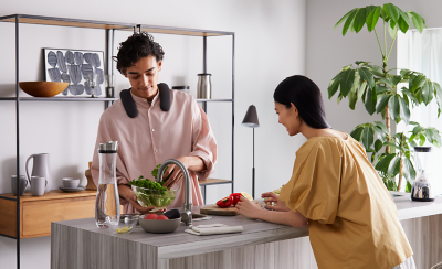 Person wearing BRAVIA Theatre U preparing food in a kitchen with person resting on countertop and ornaments behind on a display unit and large pot plant to right