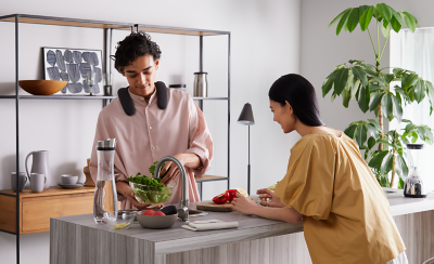 Person wearing BRAVIA Theatre U preparing food in a kitchen with person resting on countertop and ornaments behind on a display unit and large potted plant to right