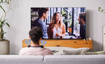 Person sitting on a sofa wearing BRAVIA Theatre U while watching movie on a large screen TV with screenshot of three people talking