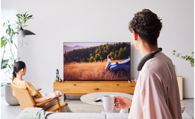Person in foreground right wearing a BRAVIA Theatre U with person left sitting in a chair, both watching TV with a screenshot of  countryside