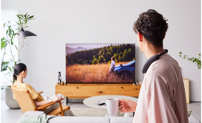 Person in foreground right wearing a BRAVIA Theatre U with person left sitting in a chair, both watching TV with a screenshot of countryside