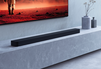 Soundbar ramping terletak di atas permukaan marmer abu-abu di bawah TV yang terpasang di dinding.