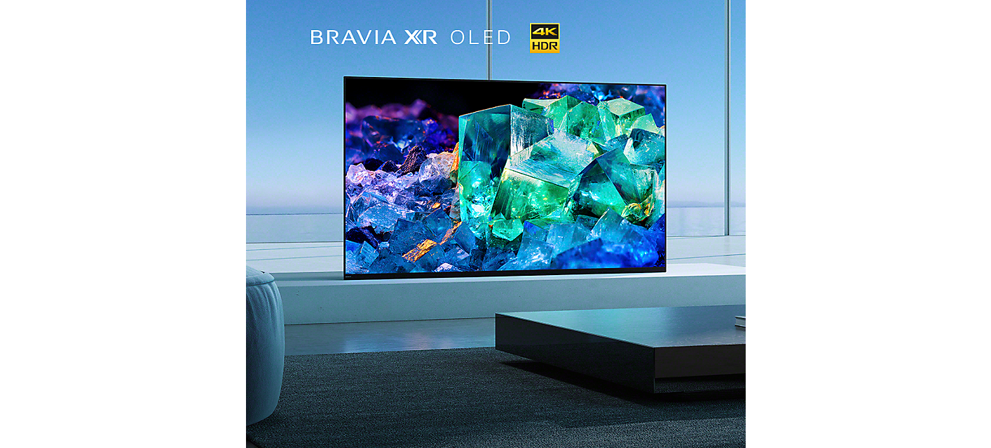 Televizor BRAVIA A95K na platformě v obývacím pokoji s knihami na stole v popředí zobrazuje modré, fialové a zelené sklo a krystaly
