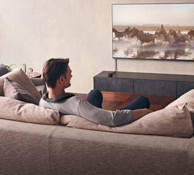 一名坐在沙發的男性，正使用 SRS-NS7 穿戴式揚聲器，並搭配 WLA-NS7 無線傳輸器，觀看壁掛電視中的影片。
