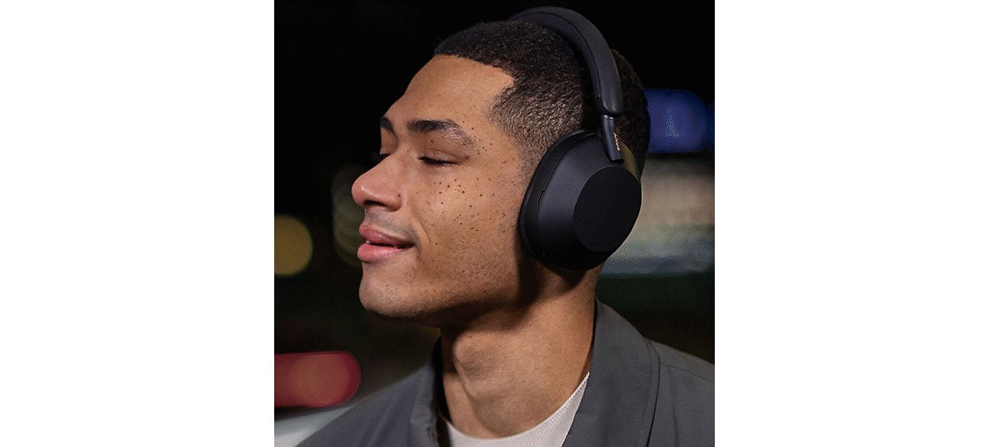 Bilde av en person som lytter til et par svarte WH-1000XM5-hodetelefoner med øynene lukket og et smil om munnen