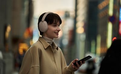 Una mujer con unos auriculares WH-CH730N de Sony en beige que sostiene un smartphone en la calle de una ciudad