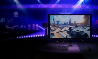 The new M10S Gaming Monitor with a first person shooter gaming graphic next to a keyboard and content camera in front of a blue and purple backround.