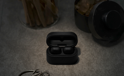  Open black earbuds case with earbuds inside, placed on a dark textured surface, surrounded by an out-of-focus jar and a cylindrical container. Moody ambiance.