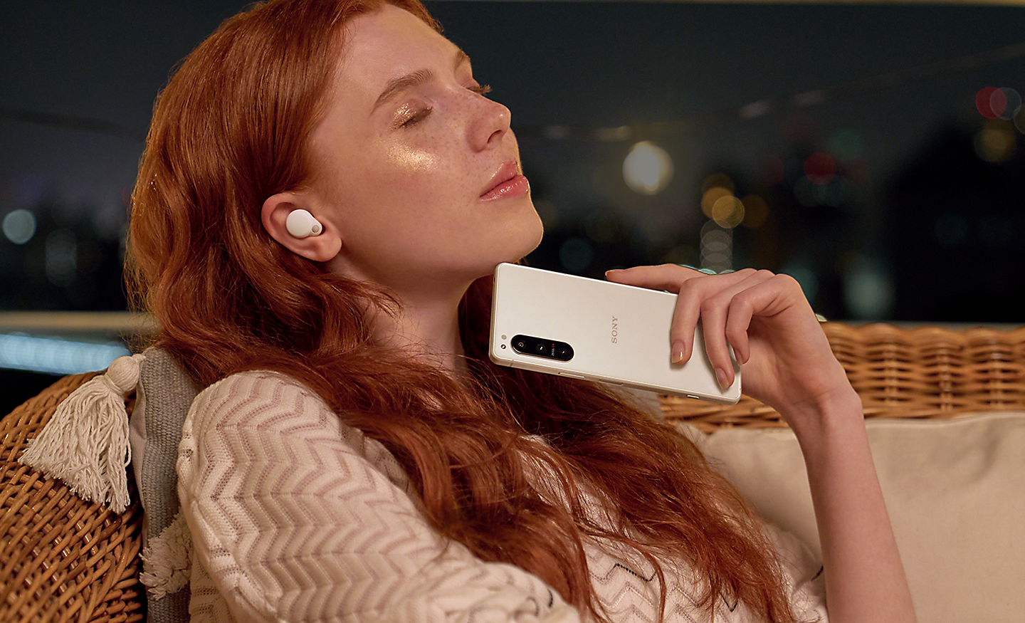 Žena se zavřenýma očima má nasazena bezdrátová sluchátka do uší a drží v ruce smartphone Xperia 5 IV