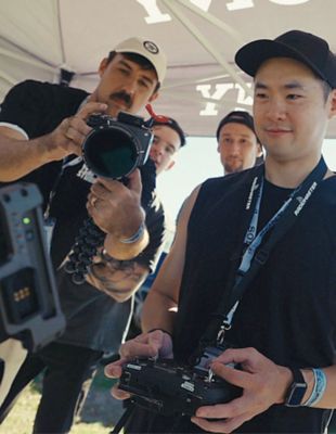 Creadores sosteniendo cámaras en un evento, uno grabando con un lente grande mientras el otro revisa el metraje en un monitor.