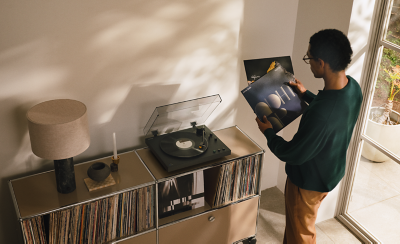 En man som väljer en vinylskiva bredvid en PS-LX5BT-skivspelare på ett skåp fyllt med skivor i ett solupplyst vardagsrum intill ett stort fönster.