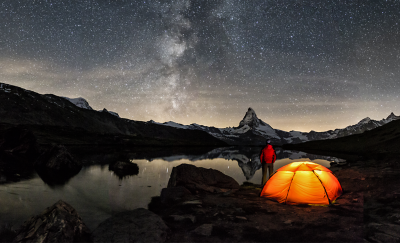Une tente orange bien éclairée sur un rivage rocheux sous un ciel nocturne clair et étoilé, avec une personne vêtue d’une veste rouge se tenant à proximité. Les montagnes enneigées se reflètent dans l’eau calme et la Voie lactée est visible dans le ciel.