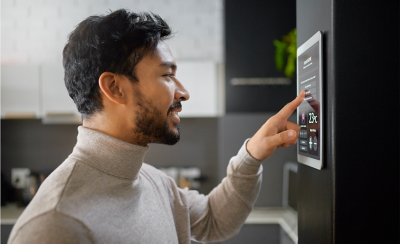 Side profile view of a person with short dark hair and beard, wearing a light-coloured turtleneck, adjusting settings on a smart home control panel mounted on a kitchen wall.