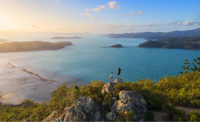 Panoramic, aerial shot of a tropical coastline at sunset, showing a vast expanse of ocean, distant mountains, scattered islands, and two people standing on a rocky outcrop, admiring the view.