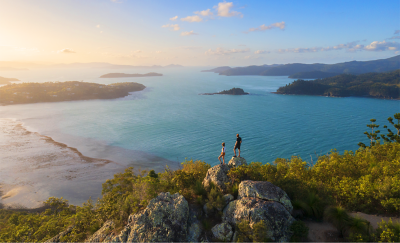 Panoramic, aerial shot of a tropical coastline at sunset, showing a vast expanse of ocean, distant mountains, scattered islands and two people standing on a rocky outcrop, admiring the view.