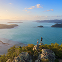 Vue aérienne panoramique d’un littoral tropical au coucher du soleil, montrant une vaste étendue d’océan, des montagnes lointaines, des îles éparses et deux personnes debout sur un affleurement rocheux, admirant la vue.