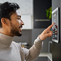 Vue de profil d’une personne aux cheveux bruns courts et portant la barbe, vêtue d’un col roulé de couleur claire, en train de régler les paramètres d’un tableau de commande de maison intelligente installé sur le mur d’une cuisine.
