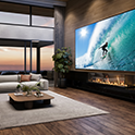 BRAVIA Projector 7 fixé au plafond montrant une superbe scène de surf sur un grand écran dans un salon luxueux, avec une cheminée moderne et une vue pittoresque sur le coucher de soleil.