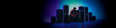 Una pared dinámica formada por los parlantes ULT POWER SOUND apilados en diferentes orientaciones verticales y horizontales. El contorno de cada unidad está delineado por brillantes anillos de luz LED azul neón sobre un fondo degradado azul y verde azulado. Una persona con una chaqueta oscura con rayas naranjas y lentes de sol está sentada en el parlante ULT TOWER MAX en el centro.