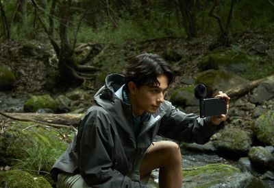Foto de una persona con una chaqueta gris resistente al agua agachada en una zona boscosa sosteniendo un smartphone con un micrófono ECM-AX10 para grabar.