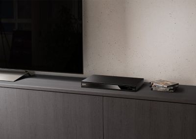 A black Blu-ray disc player is seen between a stack of blu-ray discs and a black TV.