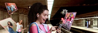 Image 102: A young woman wearing LinkBuds Fit earbuds while stood on a train platform, engaging with her phone as pop outs of social media surround her.