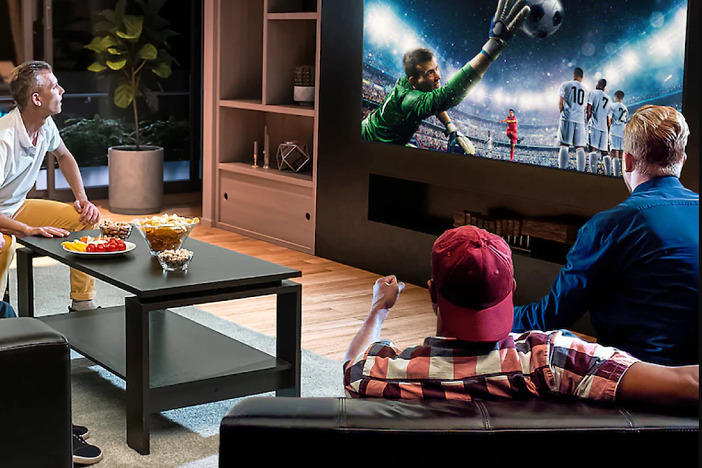Three people in a living room watch a TV displaying a goalie jumping to catch a soccer ball.